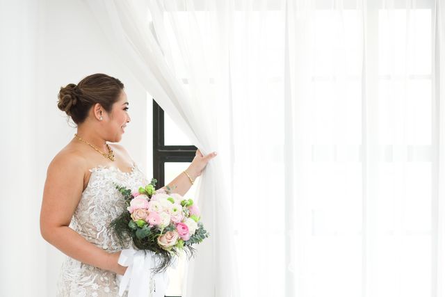 A bride in lace drawing back a curtain in soft window light